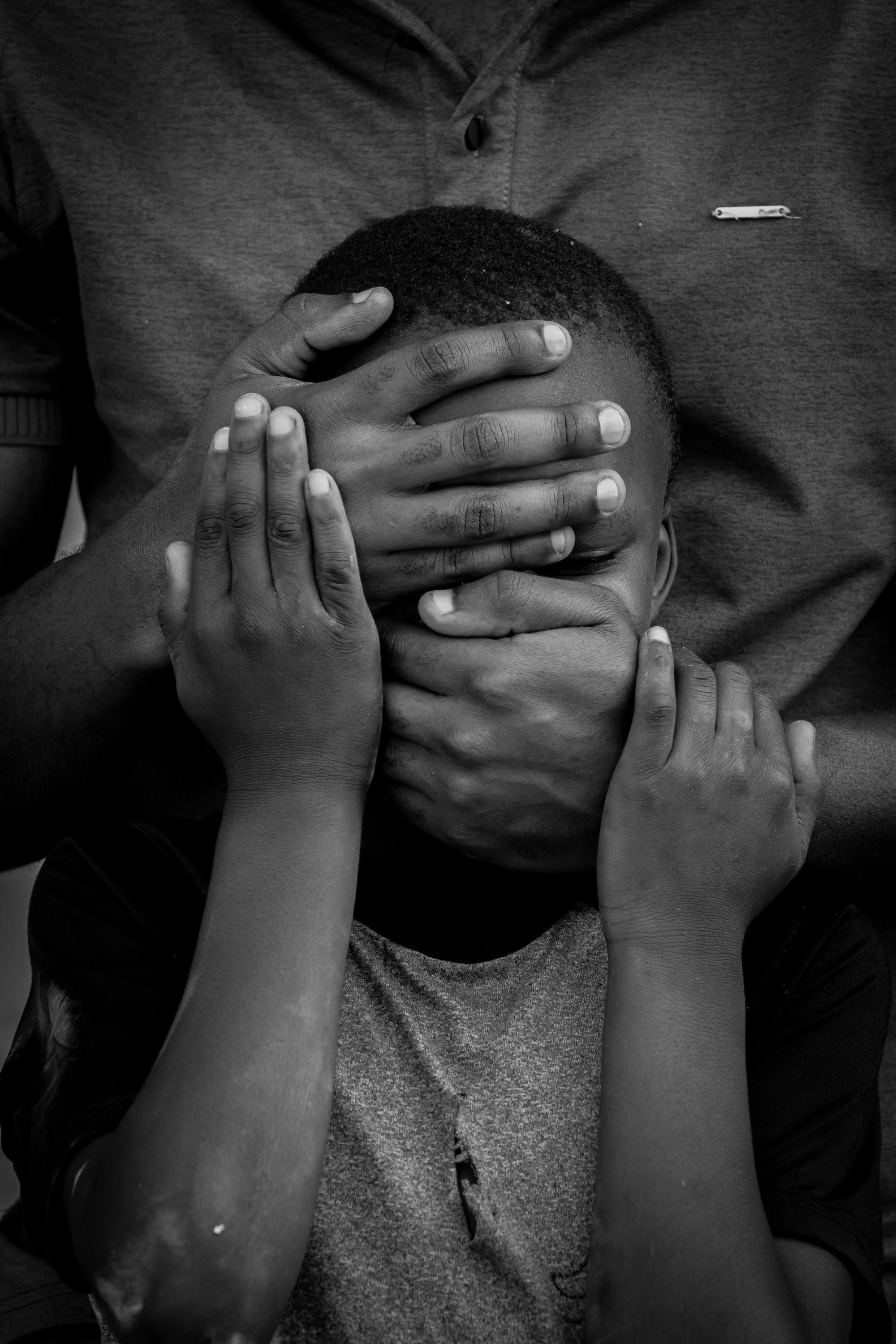 A child covering their face with both hands while an adult’s hand rests over theirs, in black and white