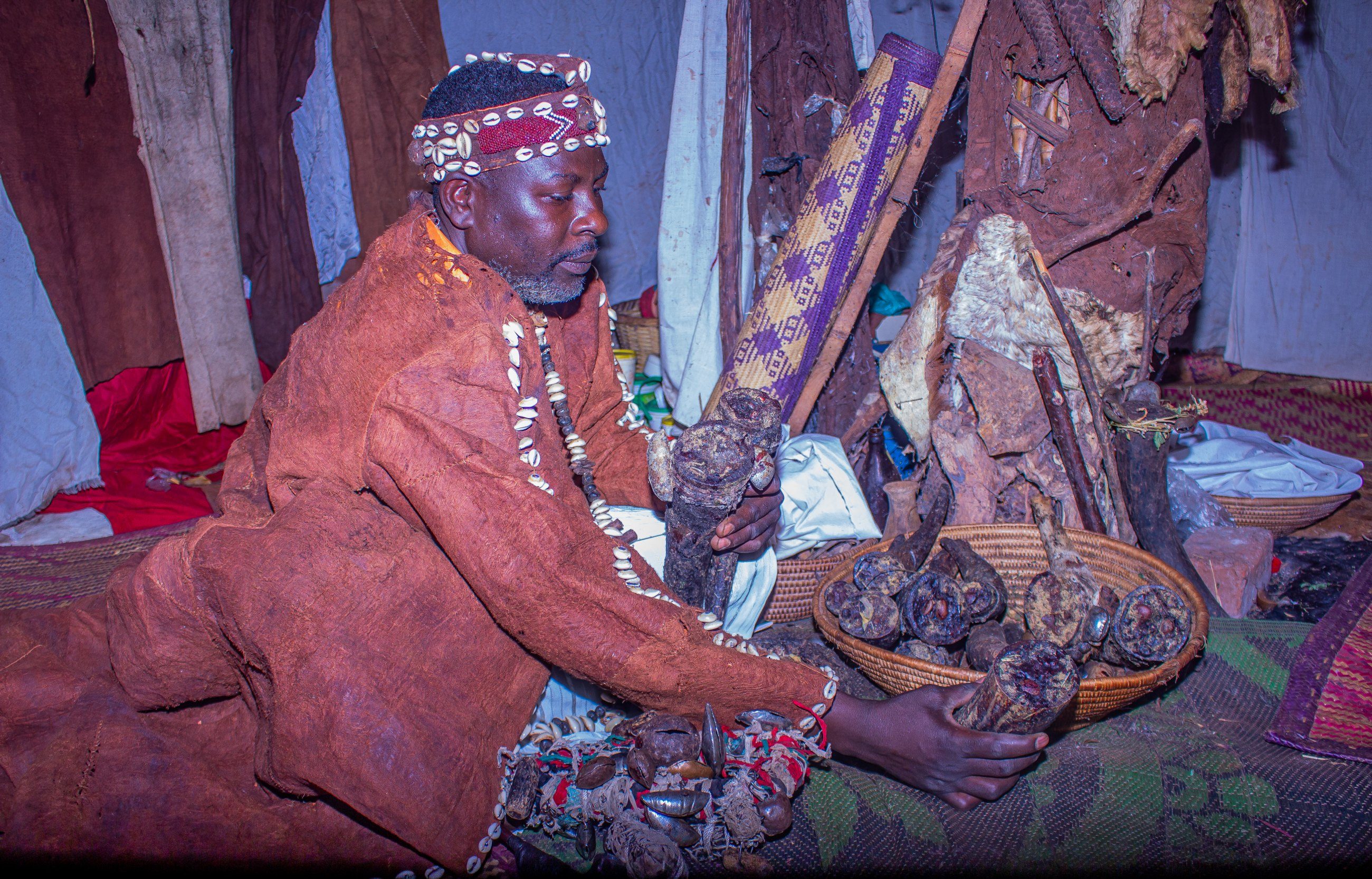 A traditional healer in a beaded headdress seated among baskets and ceremonial objects inside a shrine