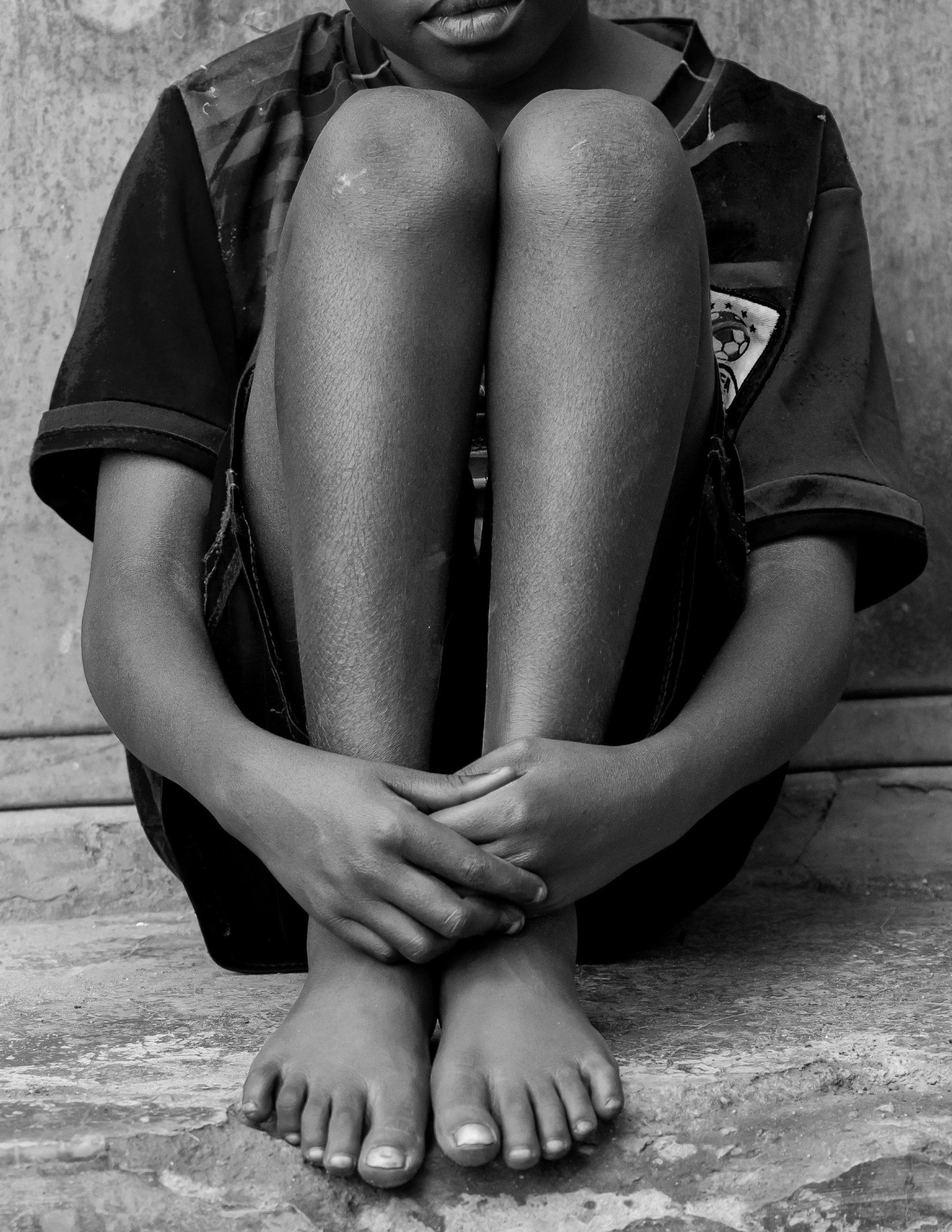 A barefoot child sitting on a concrete step with arms wrapped around their knees, in black and white