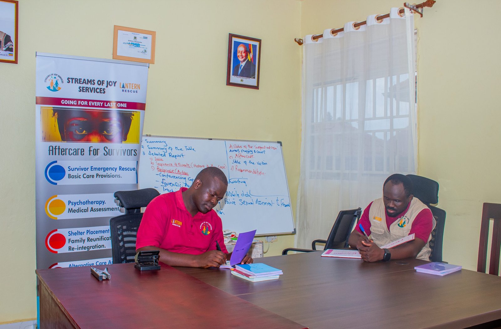 Two Streams of Joy Services caseworkers writing up records at a desk beneath a survivor aftercare chart