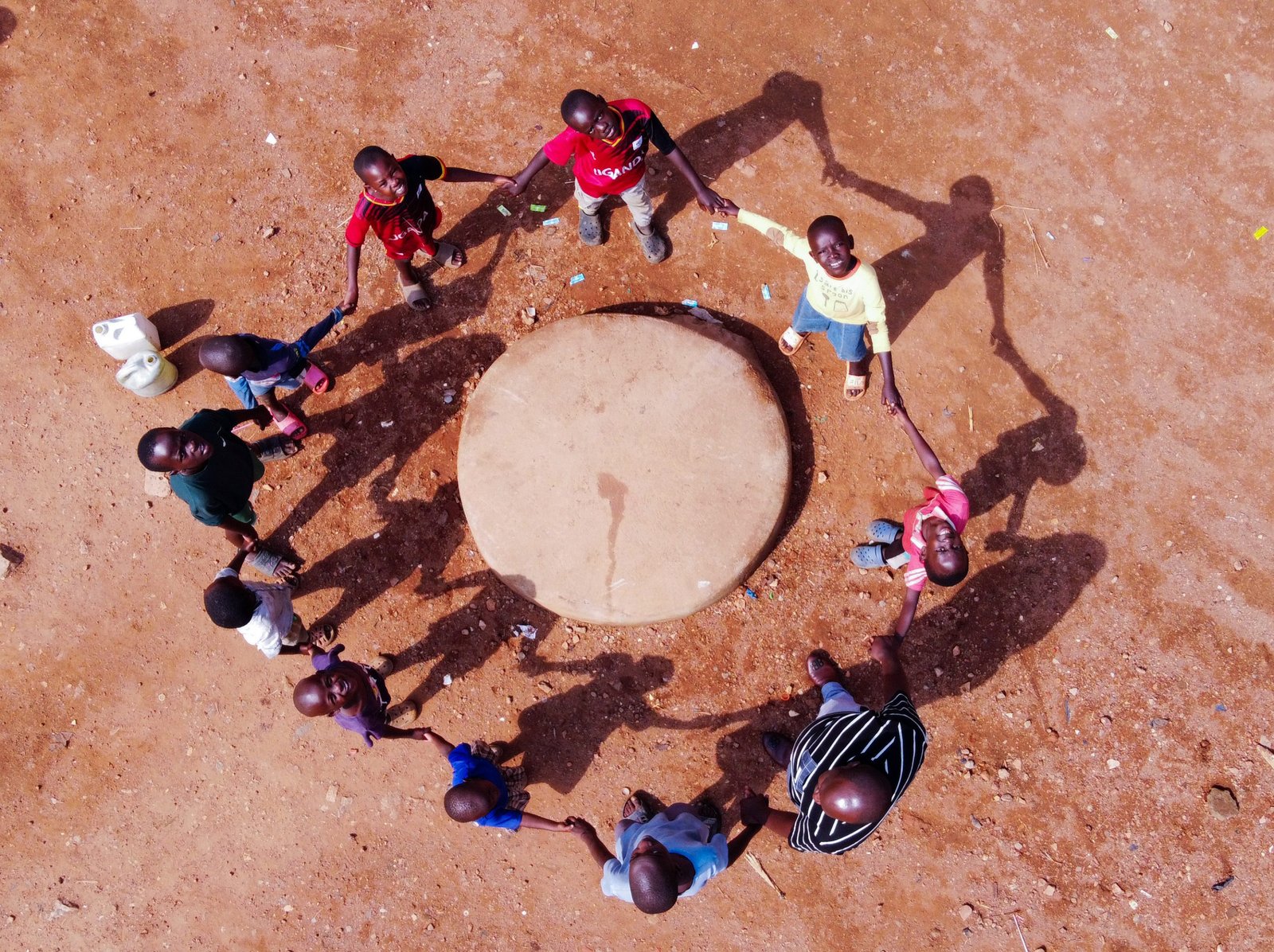 Children holding hands in a circle around a covered well on red earth, photographed from directly overhead