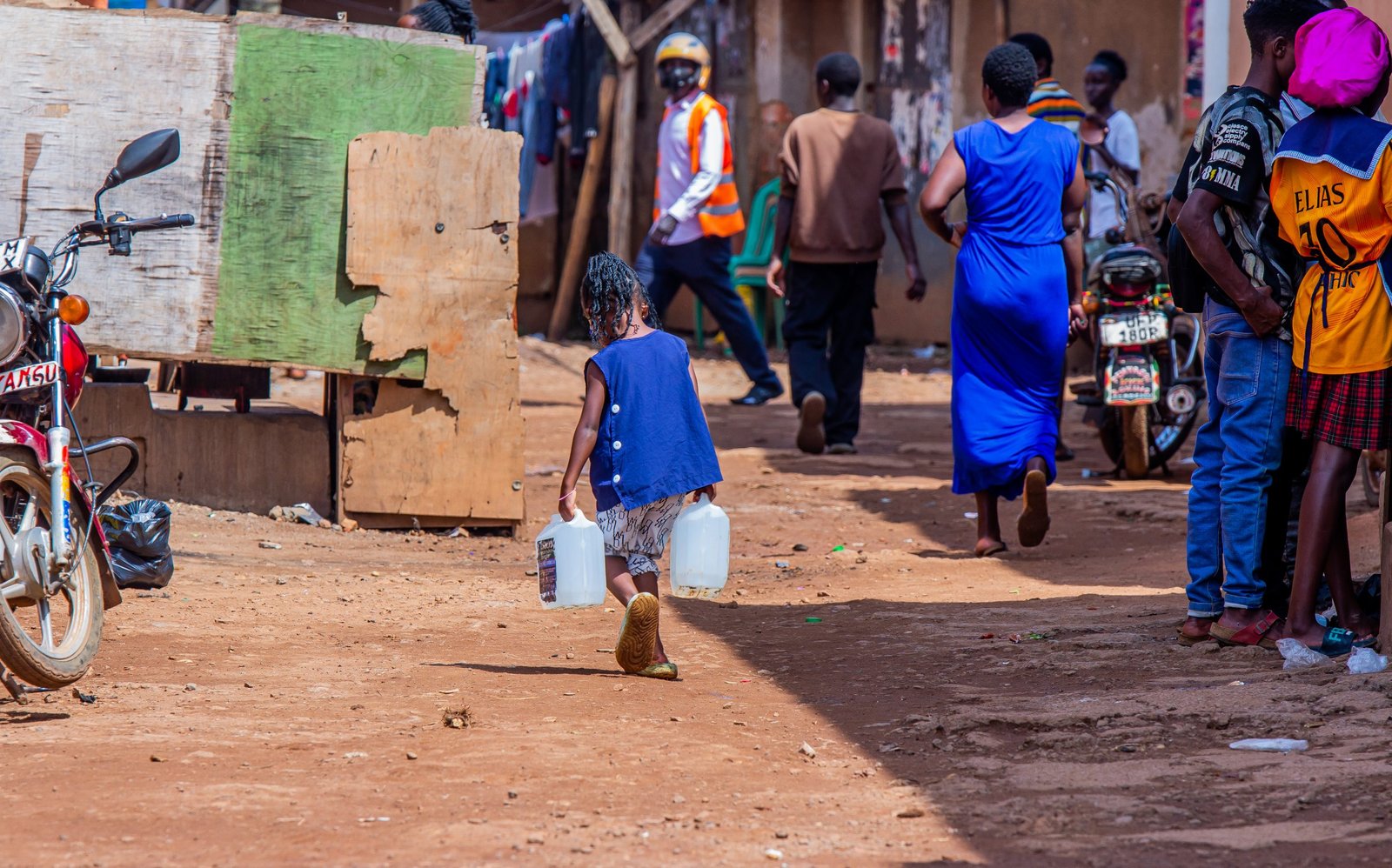 A busy neighbourhood street where a child carries water containers past shopfronts and parked motorcycles