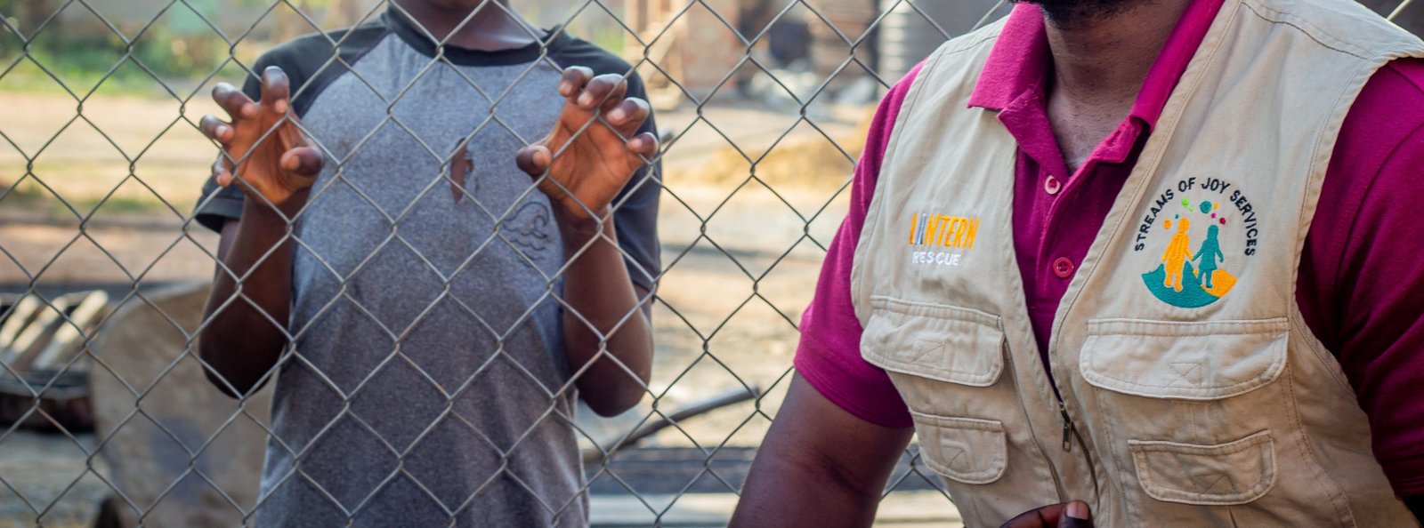 A Streams of Joy Services worker standing at a wire fence beside a child, whose face is turned away
