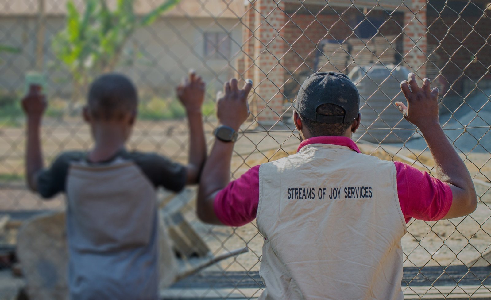 A Streams of Joy Services worker and a child standing together at a wire fence, seen from behind