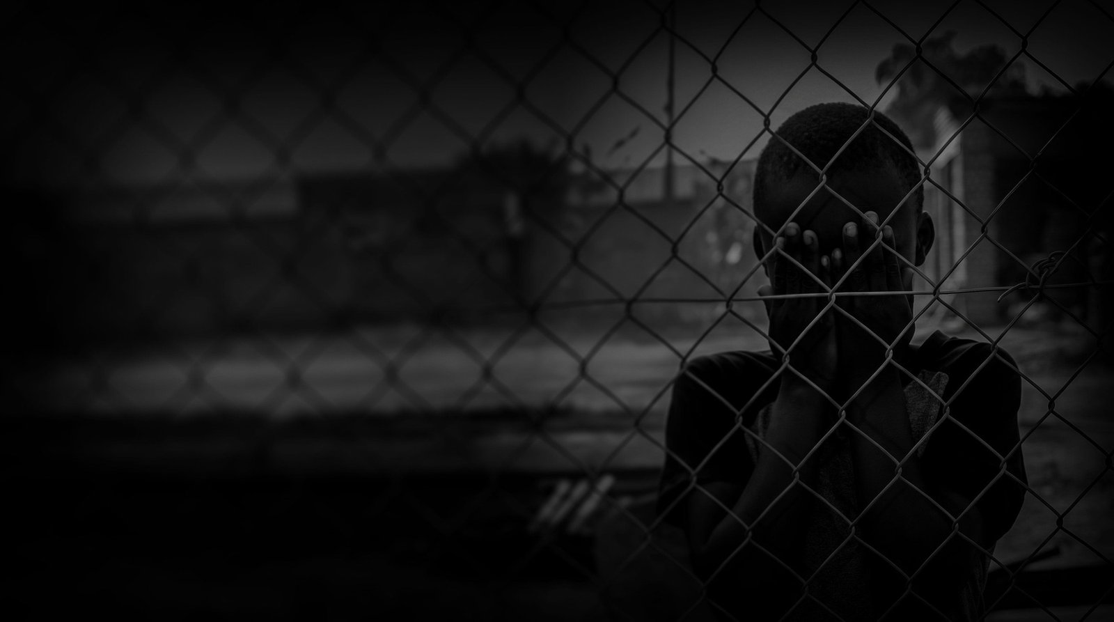 A child standing behind a chain-link fence with their hands raised to their face