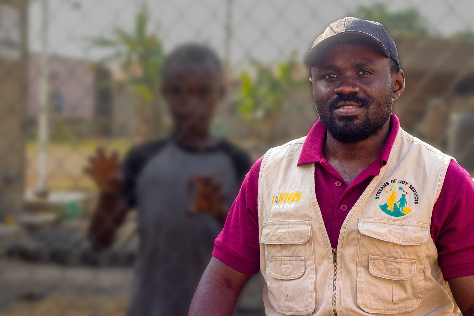 A Streams of Joy Services field officer in a branded vest, with a child standing behind him