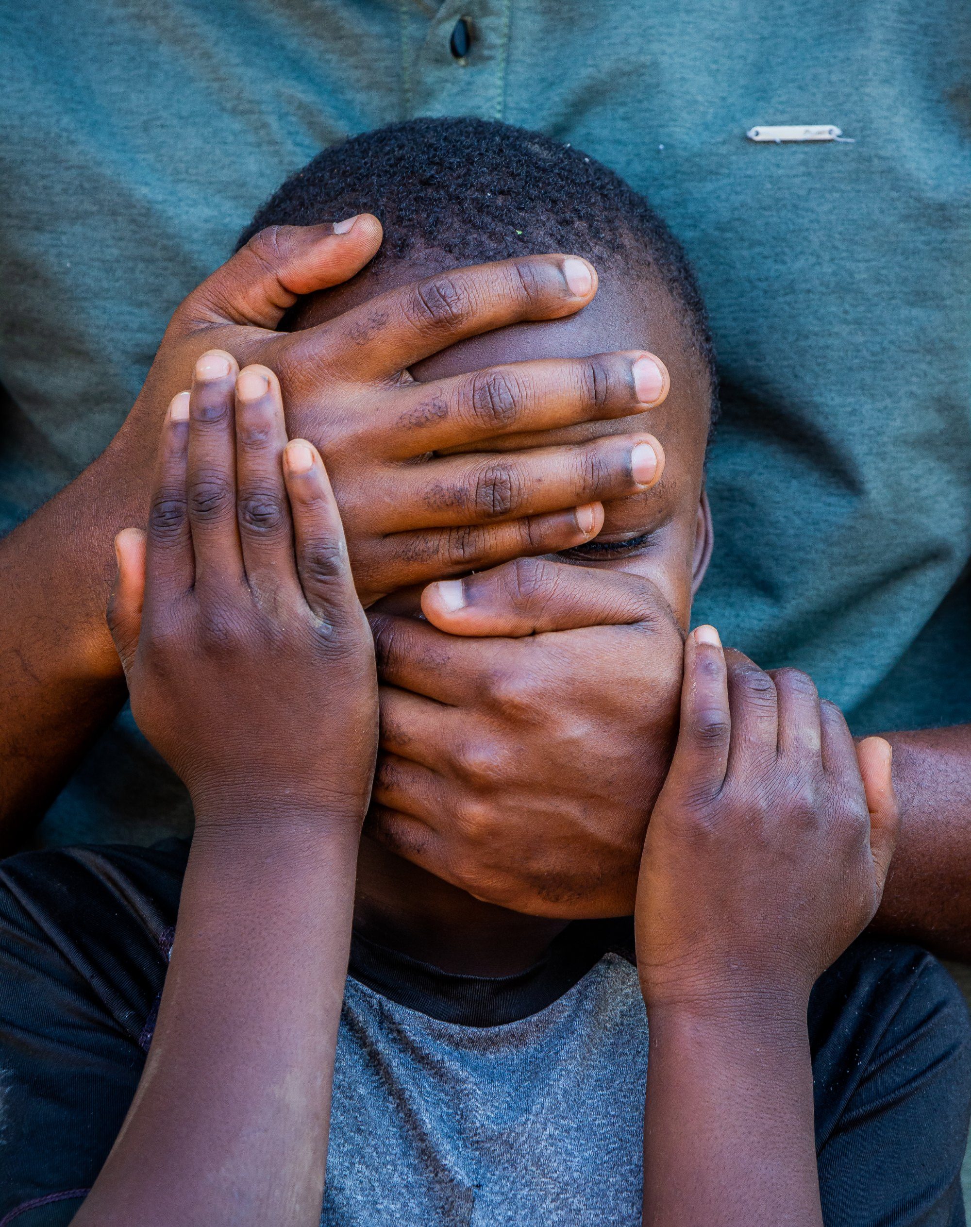 An adult hand pressed over a child's mouth while the child covers their face with both hands