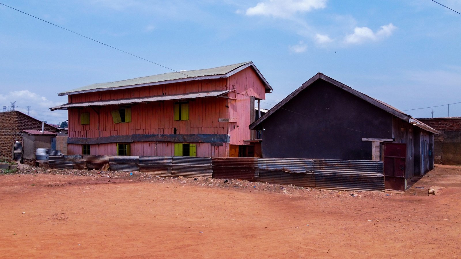 A long timber-and-iron building beside a smaller house on red earth in a Ugandan neighbourhood