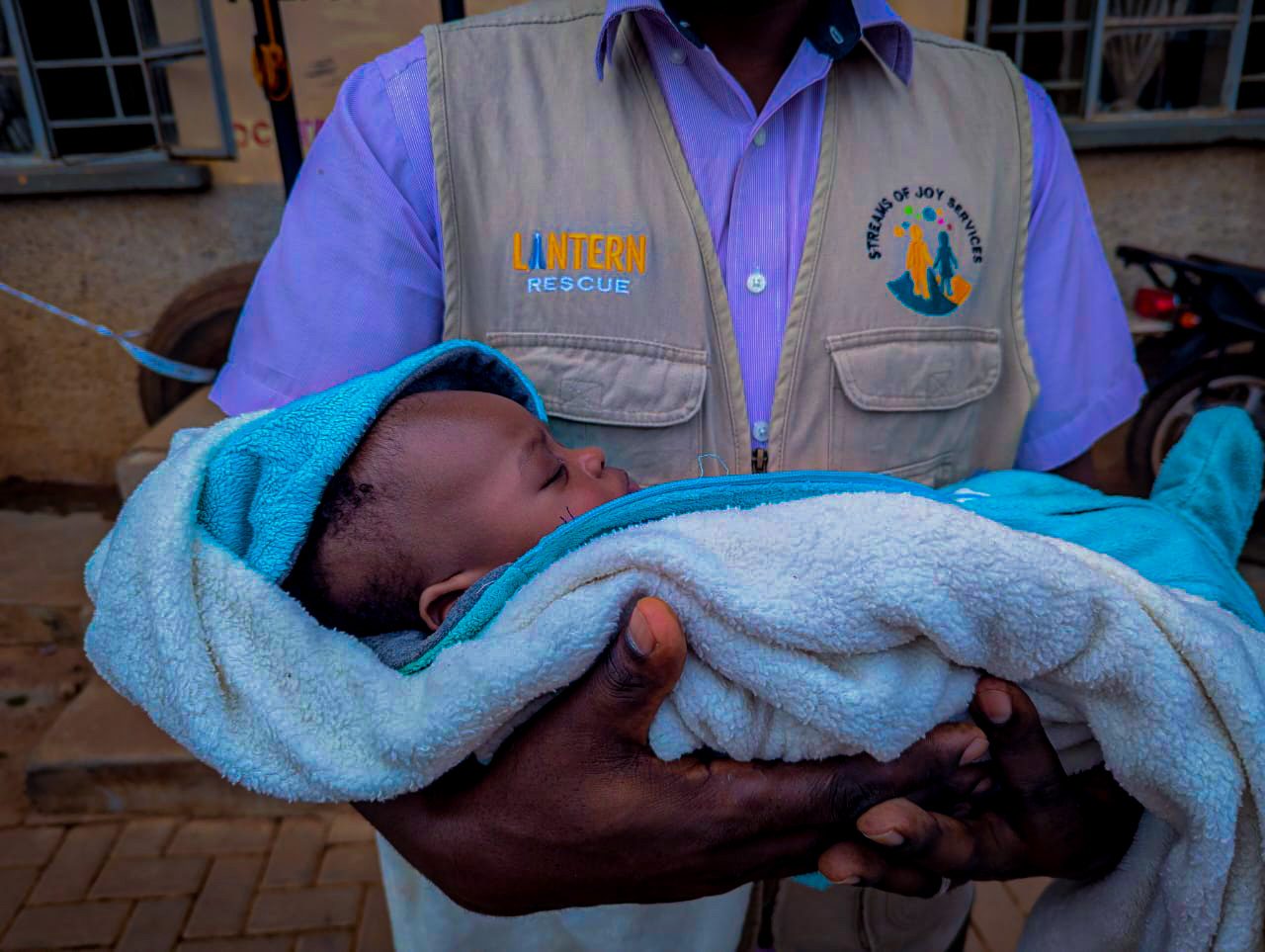 A Streams of Joy Services staff member in a branded vest cradling a sleeping baby wrapped in a blue blanket