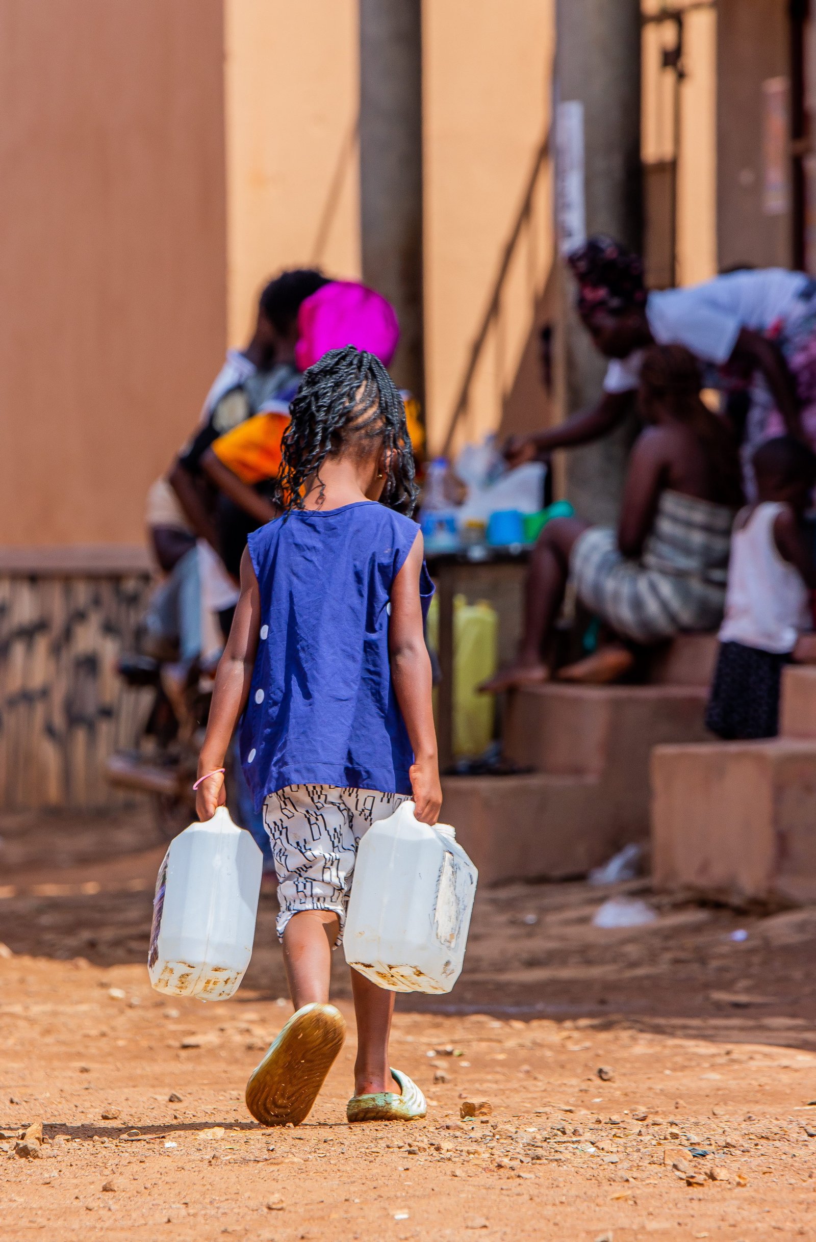 A child walking away from the camera carrying two water containers through a neighbourhood