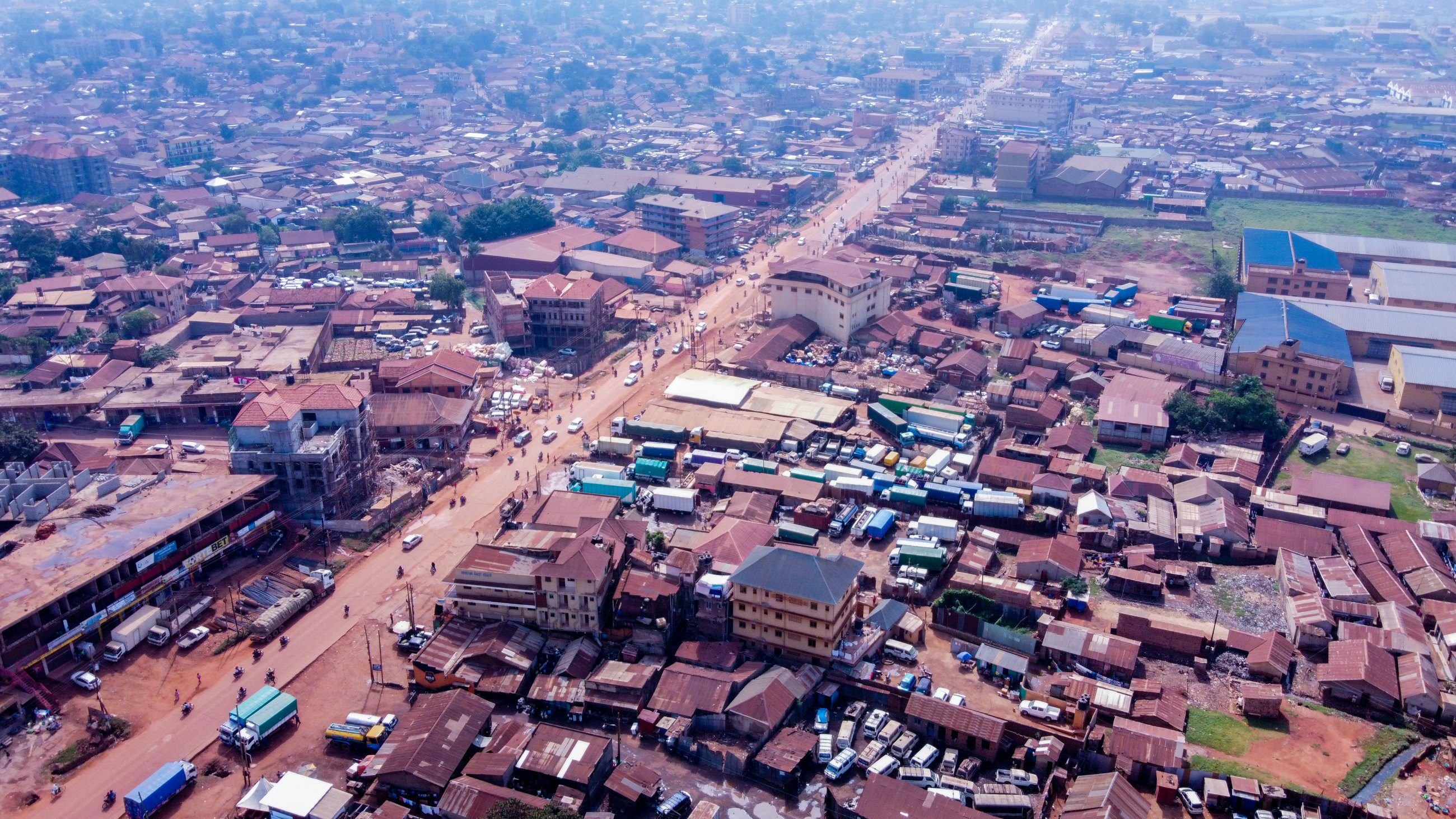 An aerial view over a Ugandan town, with a red earth main road running through densely packed rooftops toward the horizon