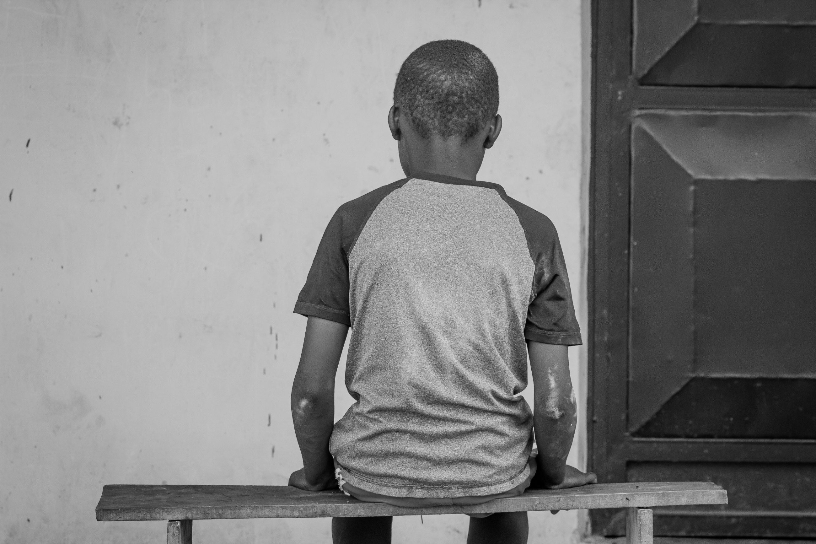 A child in a short-sleeved shirt sitting alone on a wooden bench against a plain wall, seen from behind