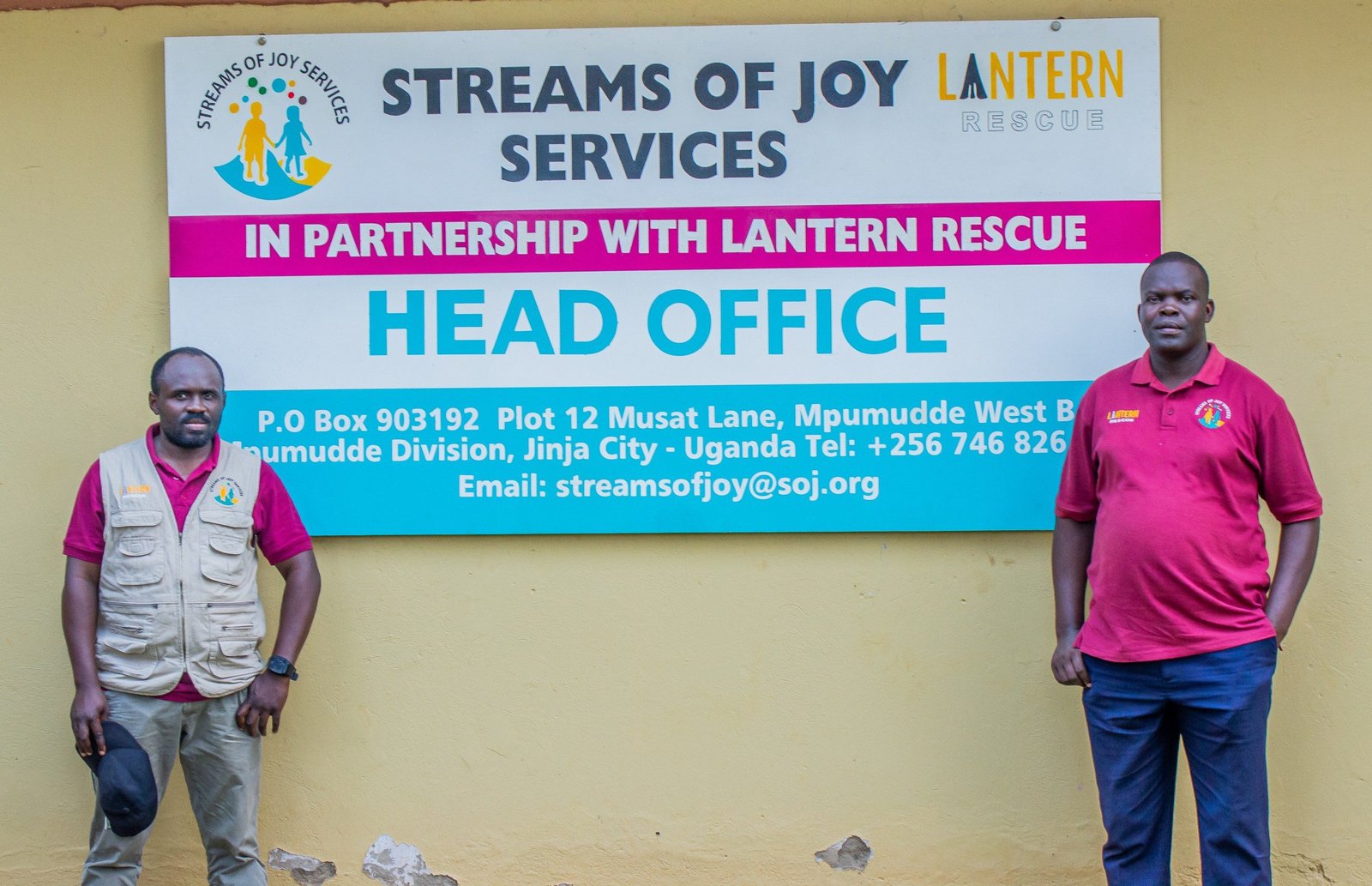 Two Streams of Joy Services staff members standing beneath the organisation's head office sign in Jinja City, Uganda